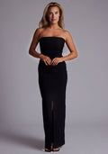 Front image of a model wearing the Vesper Alaya Bandeau Maxi Dress in Black, featuring a sleek strapless bandeau neckline, sculpting stretch fabric, and an elegant front leg split, posed against a grey studio backdrop.