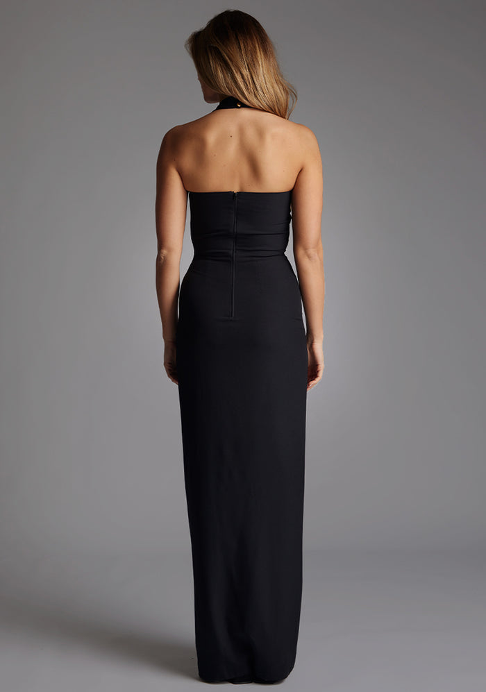 Back image of a model wearing the Vesper Amira Halter Neck Maxi Dress in Black, featuring an open upper back and a concealed centre-back zip, photographed against a grey backdrop.