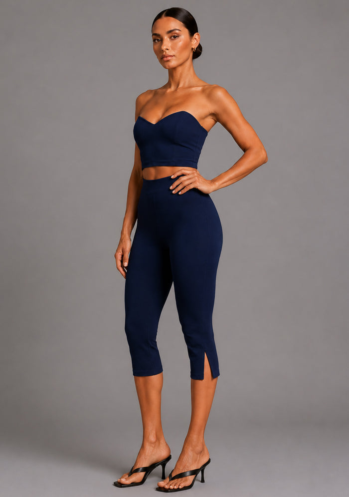 Side front image of a model wearing the VSPR Benedetta Strapless Navy Crop Top with Capri Navy Trousers in navy, showcasing the sculpted strapless bodice, clean semi-V neckline, and coordinating tailored trousers against a grey studio backdrop.