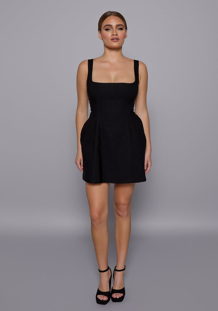 Model wearing the VSPR Camille Black Mini Dress with a square neckline and wide straps, styled with a soft A-line silhouette, standing against a minimalist grey backdrop.