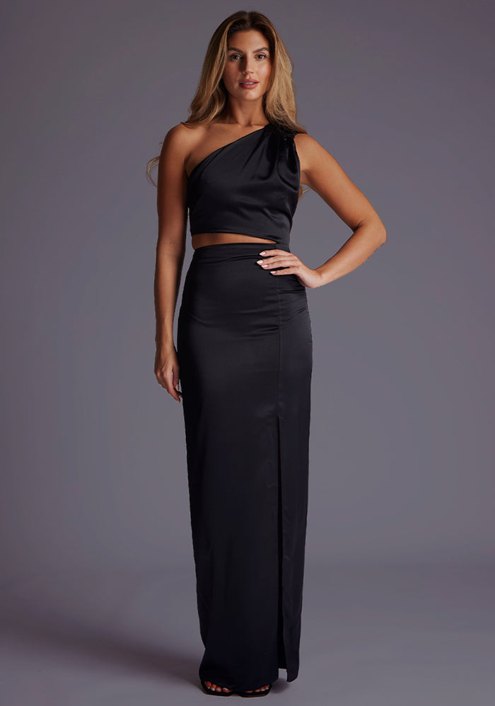 Front image of a blonde model wearing a black maxi dress with a alluring asymmetric one-shoulder design, subtle side cut-out and flattering wrap design skirt. The design featured is the Vesper Candace One-Shoulder Satin Maxi Dress in Black