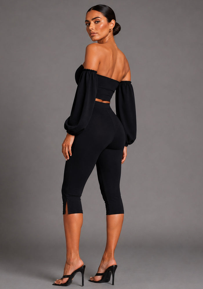Side back image of a model wearing the VSPR Belle Sweetheart Neckline Puff Sleeve Black Top highlighting the structured puff sleeves, smooth stretch fabric, and flattering body-hugging shape, The design paired with Capri Black Cropped Trousers, standing against a grey studio backdrop.