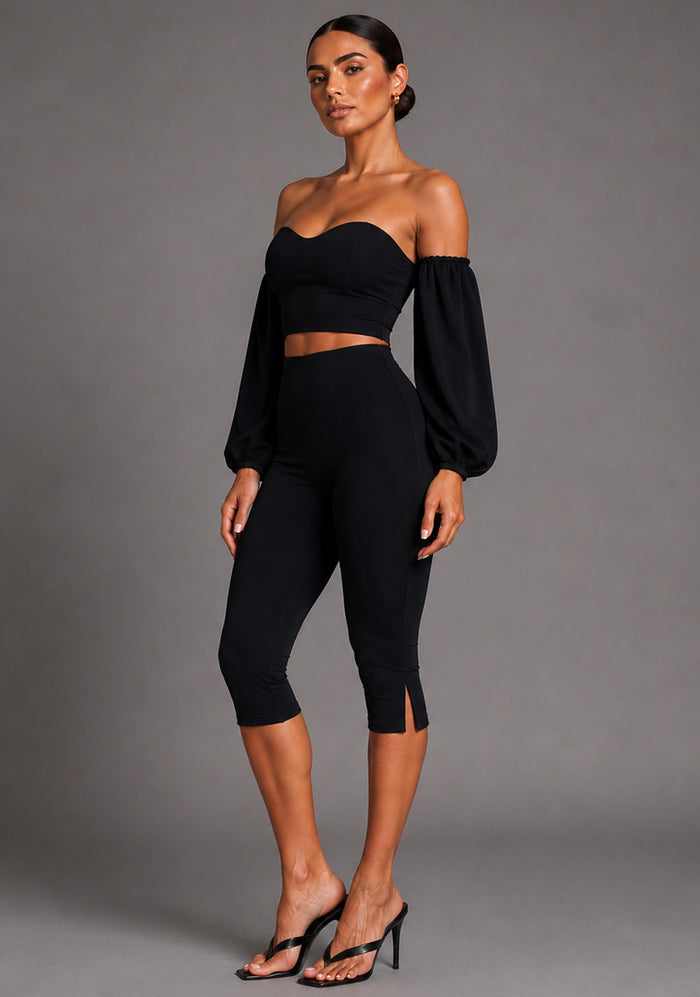 Side front image of a model wearing the VSPR Capri Cropped Pants in Black showcasing the sleek silhouette, flattering high-rise design and subtle side hem split detail. The design paired with Capri Black Cropped Trousers, against a grey studio backdrop
