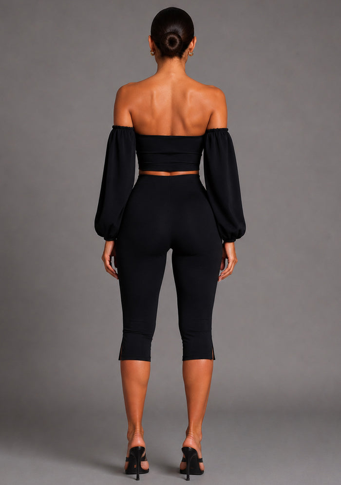 Back image of a model wearing the VSPR Belle Sweetheart Neckline Puff Sleeve Black Top featuring dramatic sleeve detailing, a sleek fitted design, and a polished silhouette. The design paired with Capri Black Cropped Trousers. posed against a grey studio backdrop.